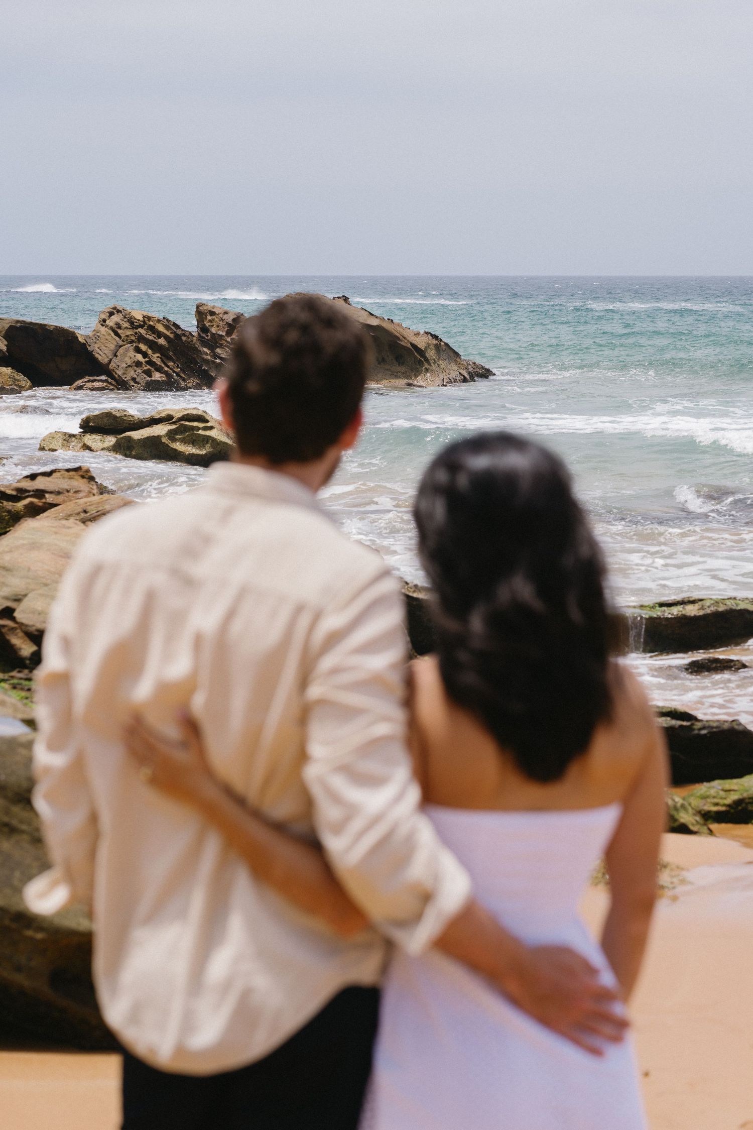 Couple embracing while looking out at the ocean waves and rocky coastline.