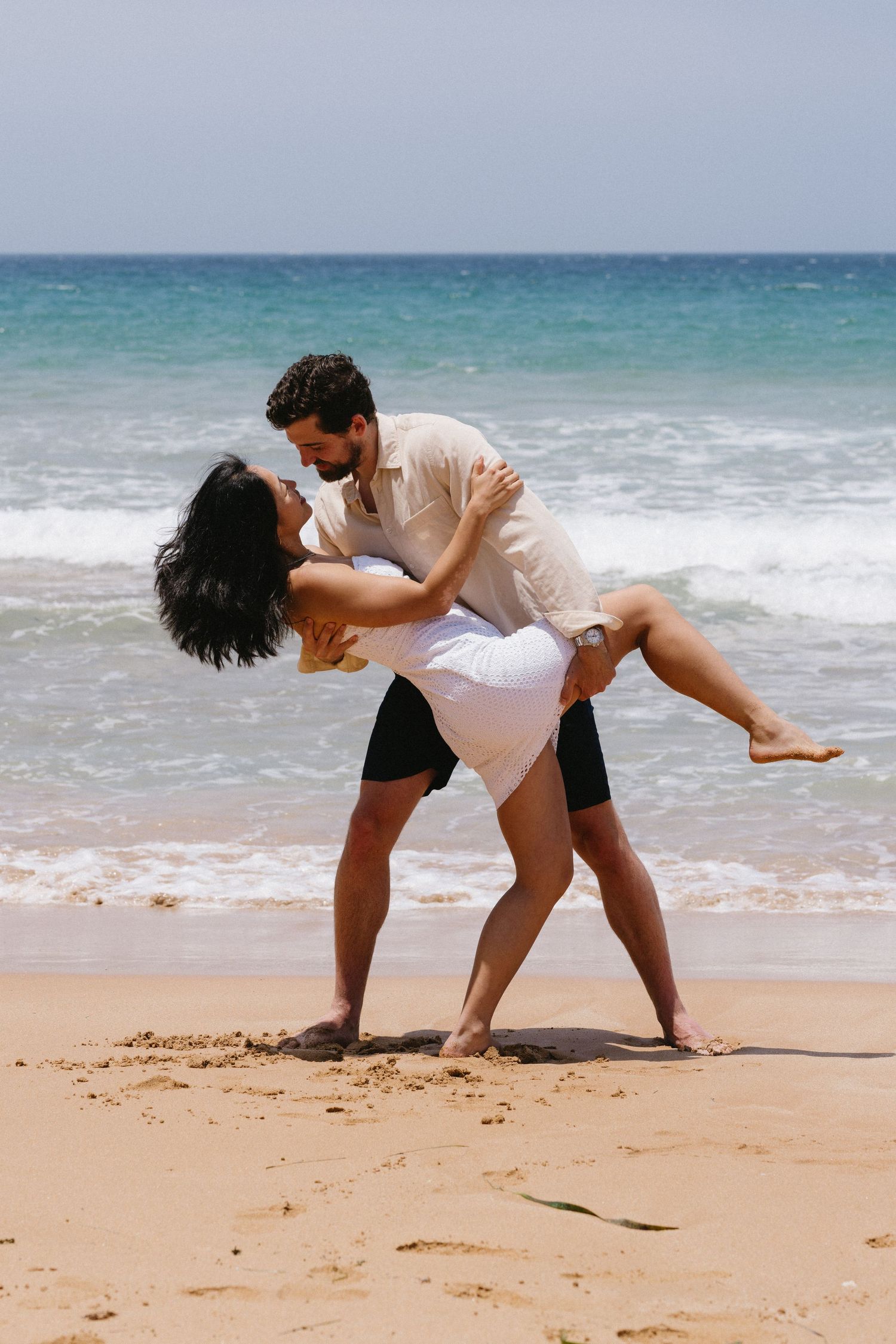 Playful couple dancing and embracing on sandy beach with turquoise ocean waters.