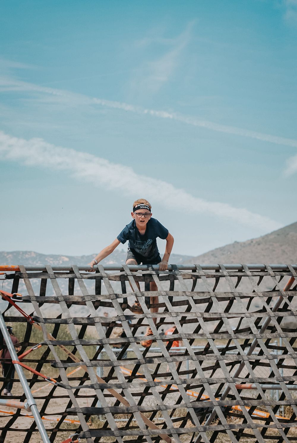 A young boy wearing sunglasses climbs a wooden lattice obstacle outdoors under a blue sky.