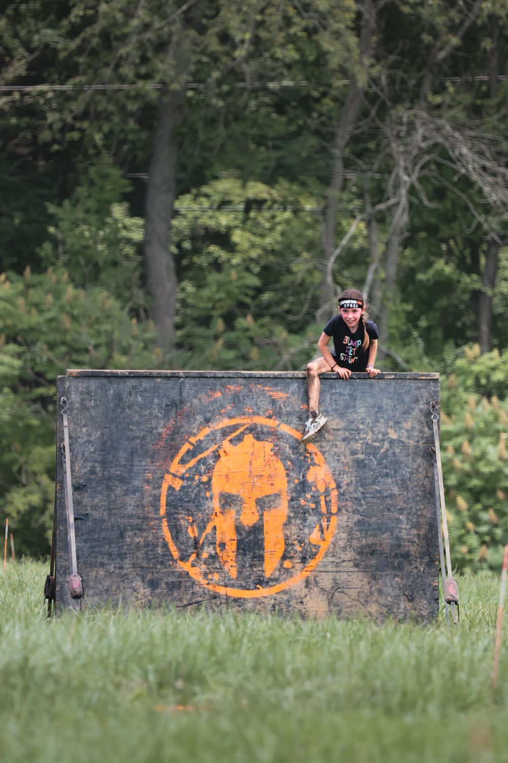 Person climbing over a Spartan Race obstacle wall with orange logo in a grassy outdoor course setting.