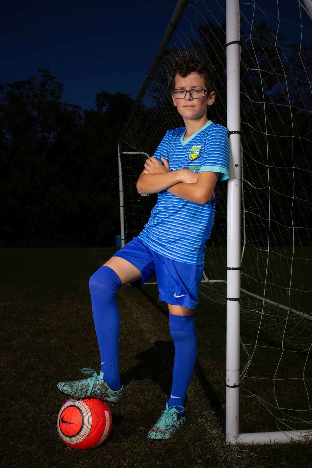 Soccer player in blue striped uniform poses with red ball by goal post during evening photo shoot.