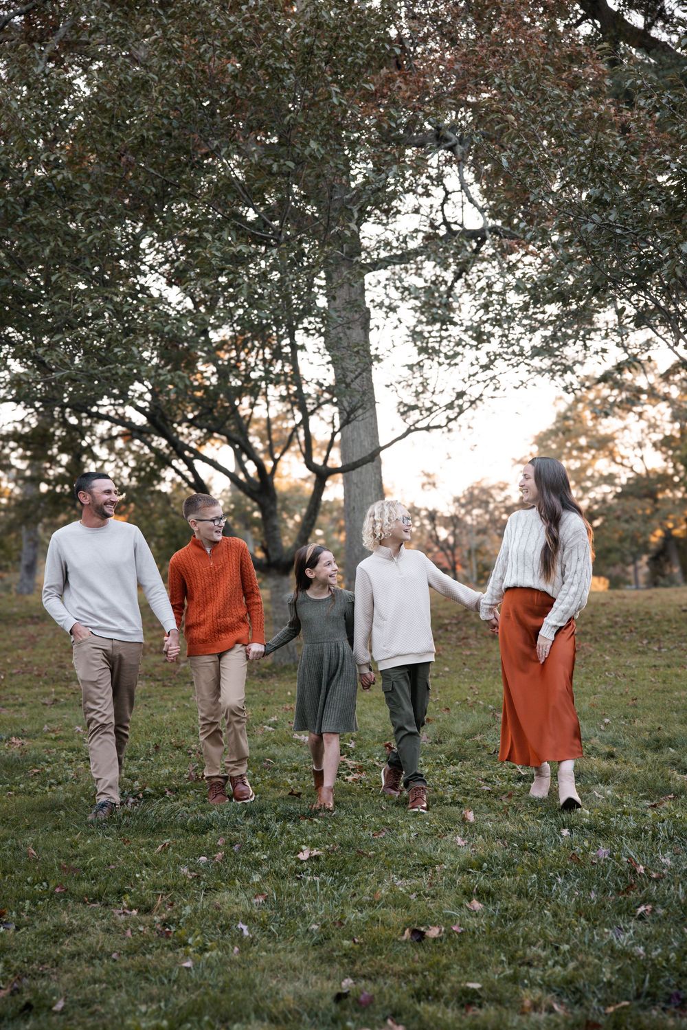 Family of five holding hands walking in a park during autumn, wearing earth-toned outfits with trees in the background.