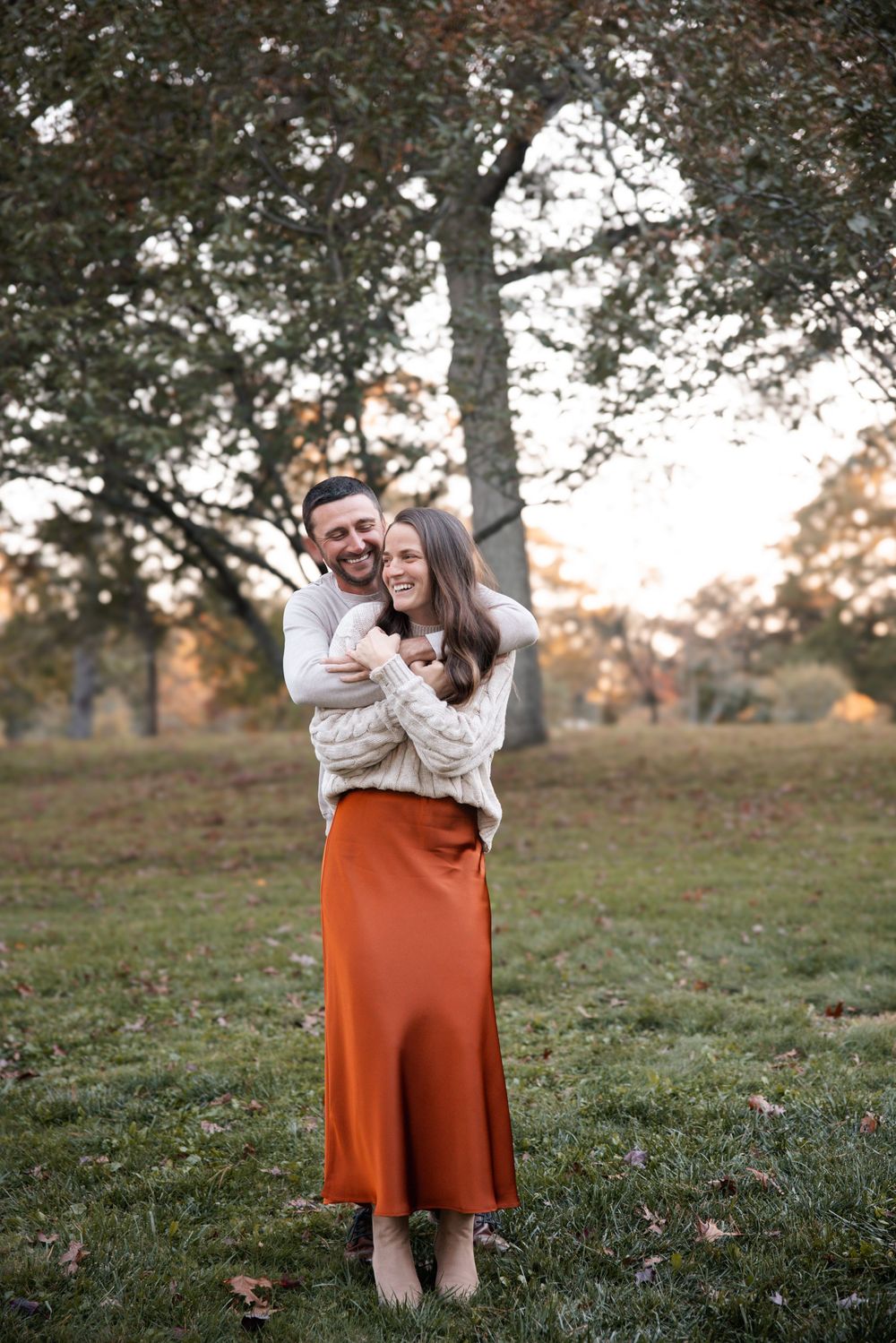 Couple embracing and laughing in a park during golden hour, woman in rust satin skirt and cream sweater.