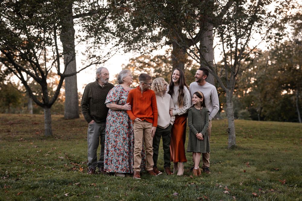 A multigenerational family poses together outdoors among autumn trees during a warm golden hour family portrait session.