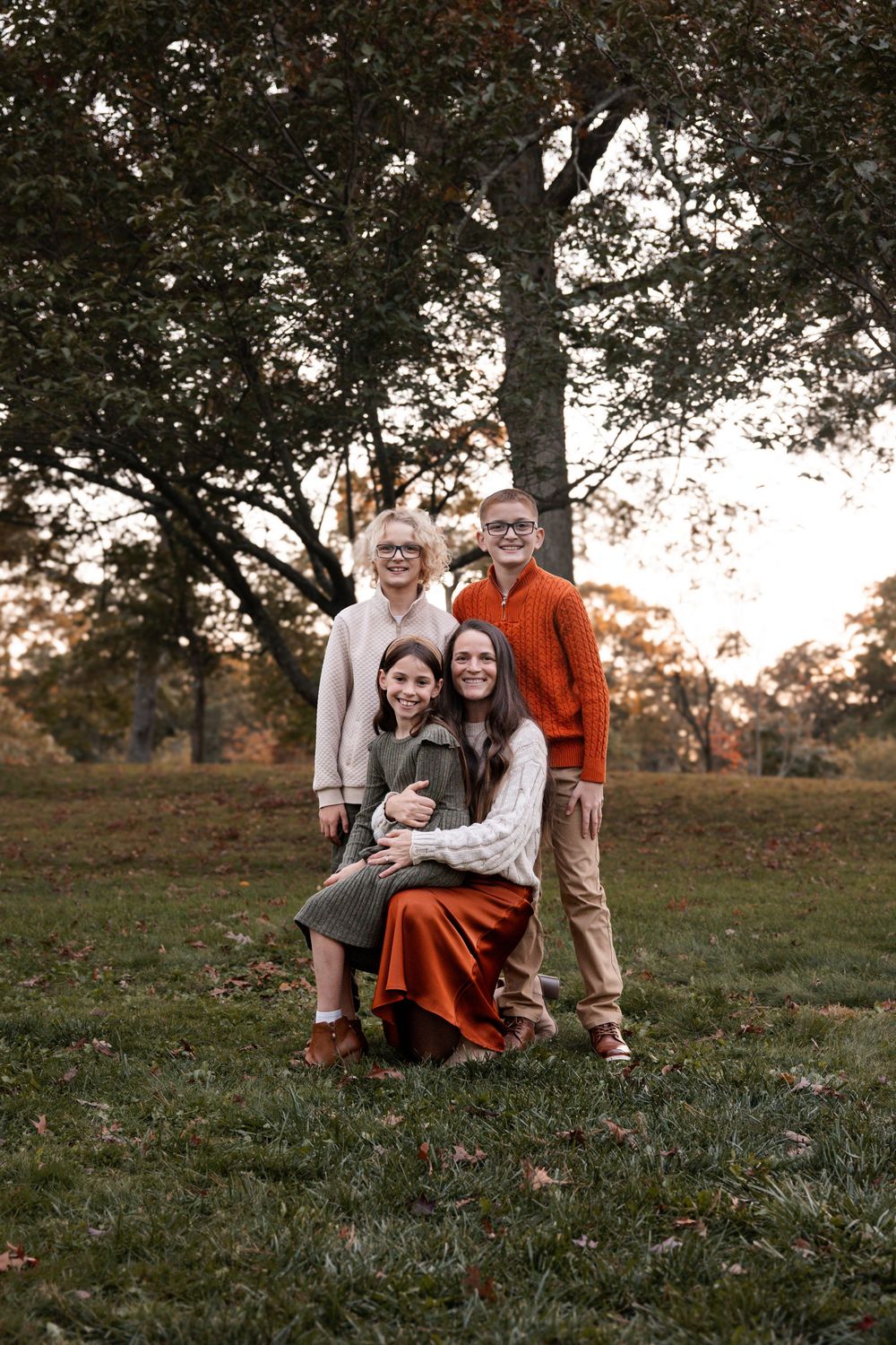 Family of four posing outdoors in autumn, smiling among fall foliage and golden trees at sunset.