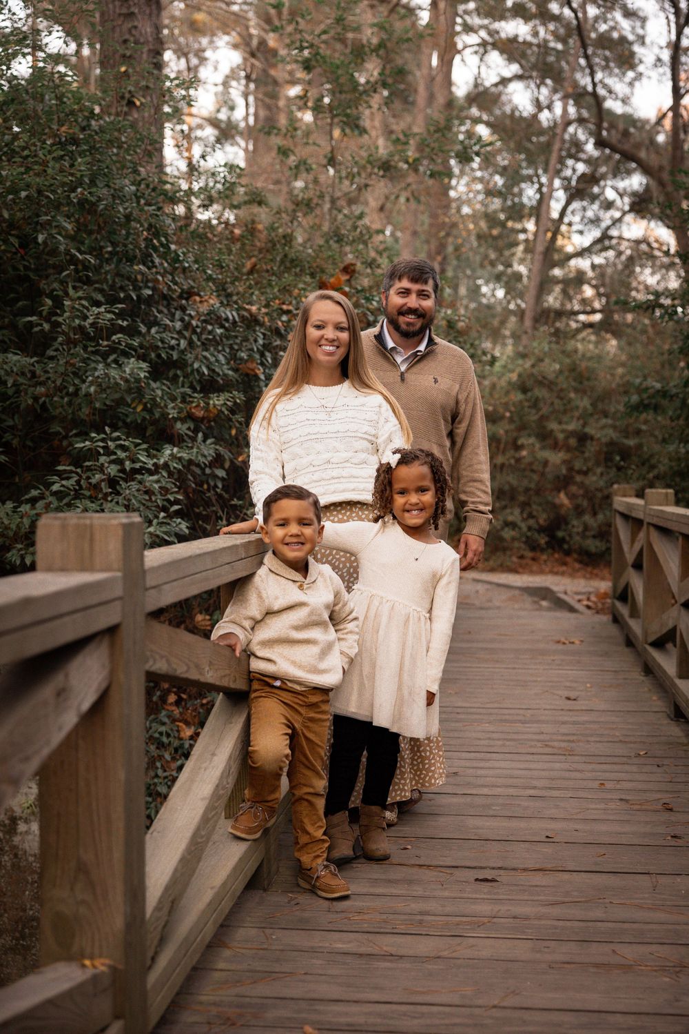 A family poses together on a wooden boardwalk surrounded by trees and greenery during an outdoor photo session.