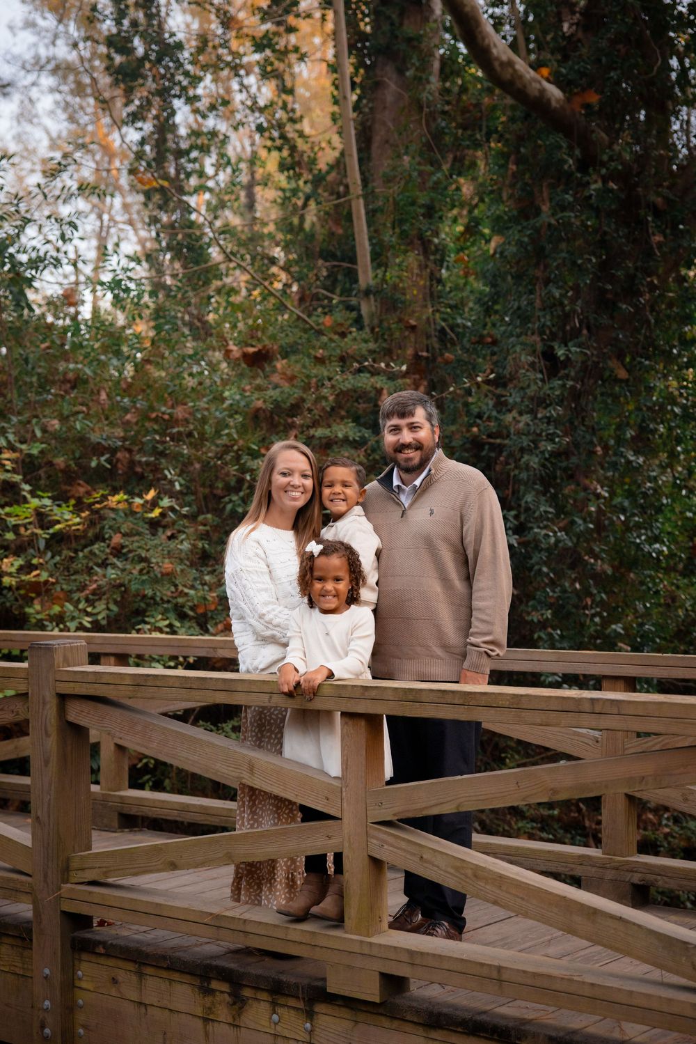 A family shares a tender moment together on a wooden bridge surrounded by autumn foliage in a park setting.