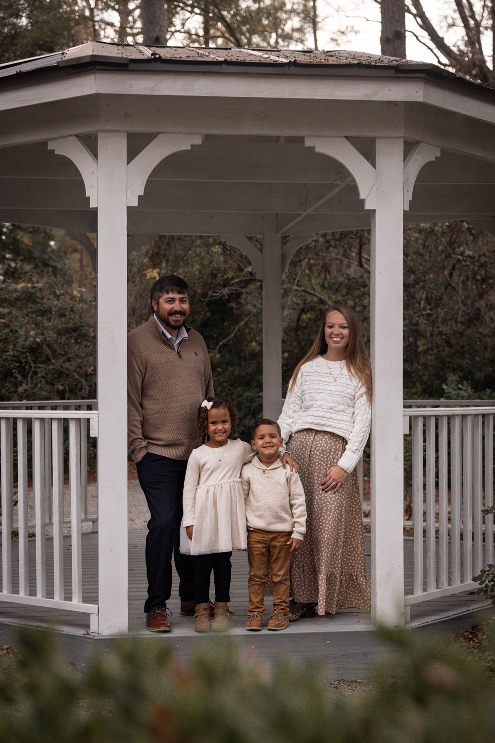 A family poses together in a white gazebo surrounded by trees and greenery during a photo session.
