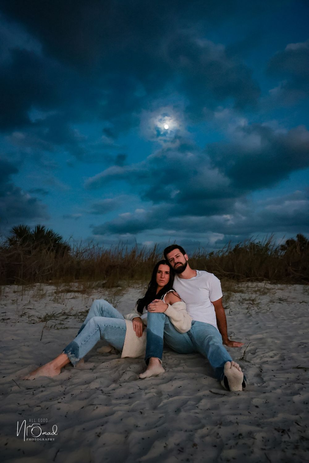 A couple relaxes on a sandy beach under dramatic moonlit clouds in casual attire.