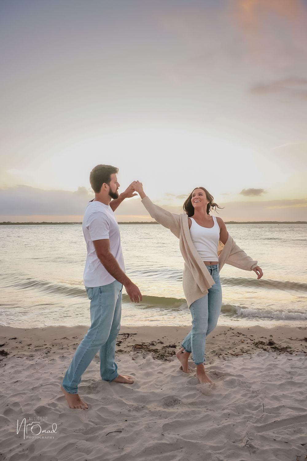 A couple playfully dances together on a beach at sunset while wearing casual white tops and jeans.