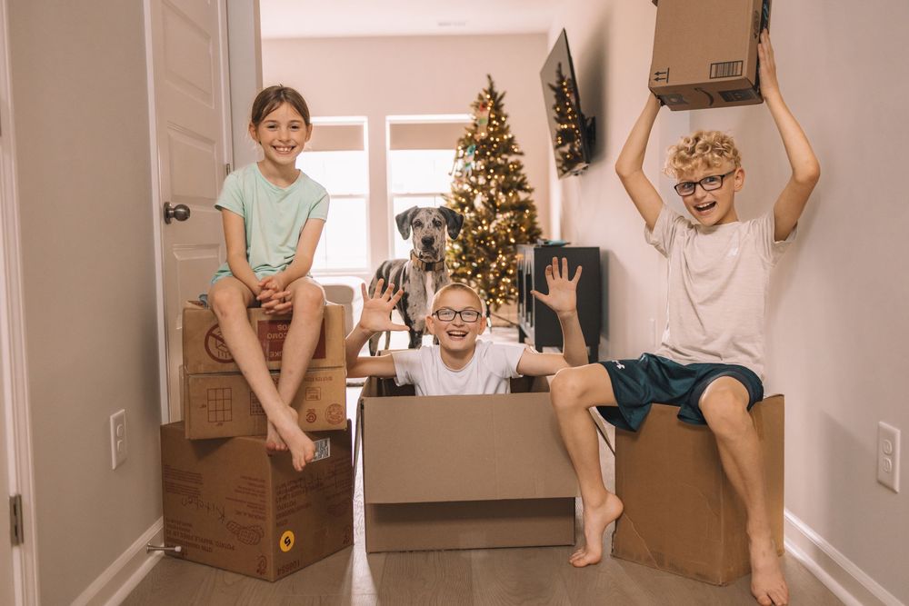 Three kids playing with cardboard boxes near a decorated Christmas tree in a bright home hallway.