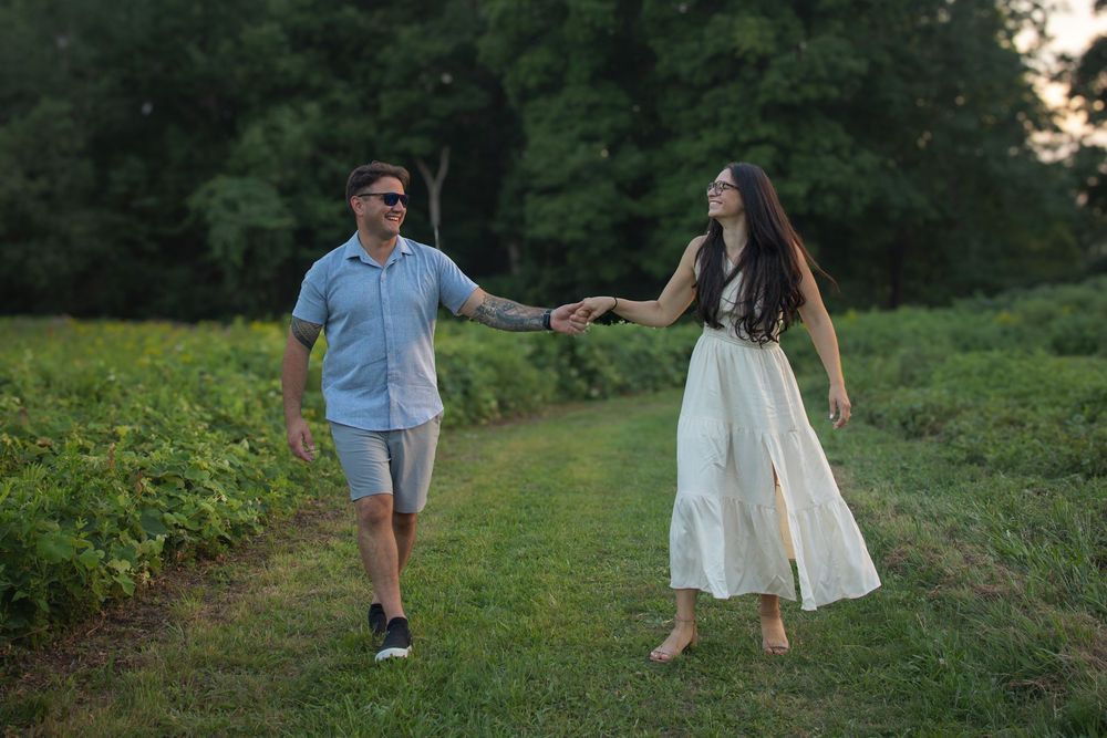 Couple holding hands walking on a green grass path surrounded by lush trees in a romantic outdoor setting.