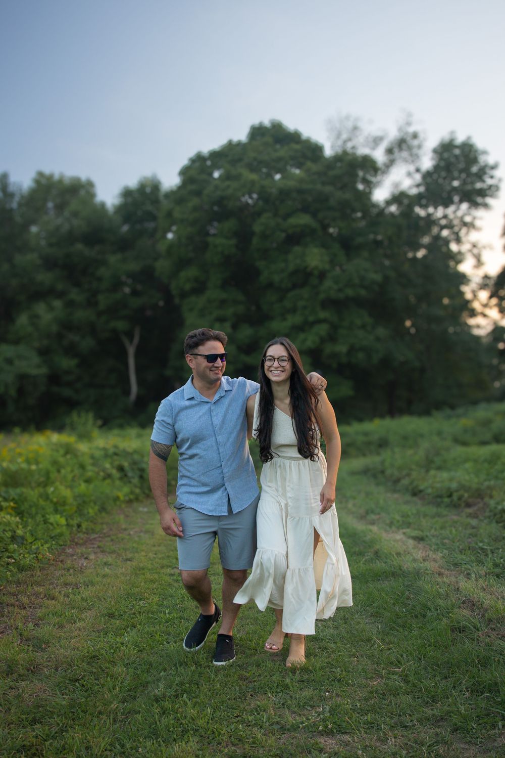 A couple walks hand in hand through a lush green field at dusk, smiling and dressed in casual summer attire.