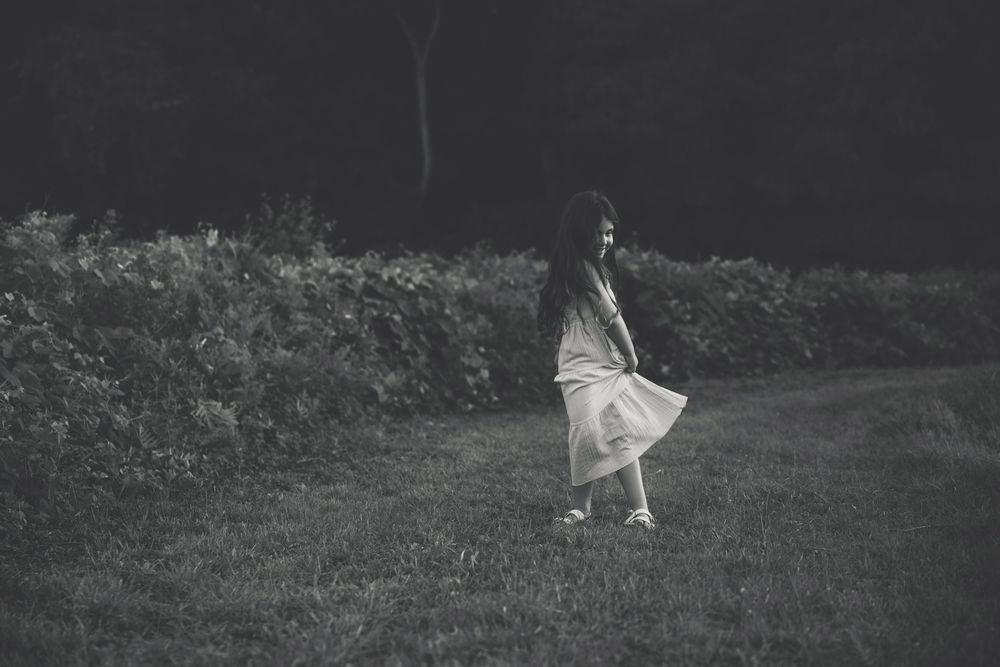 Black and white photo of a young woman in a flowing dress walking on grass near dark hedges at dusk.