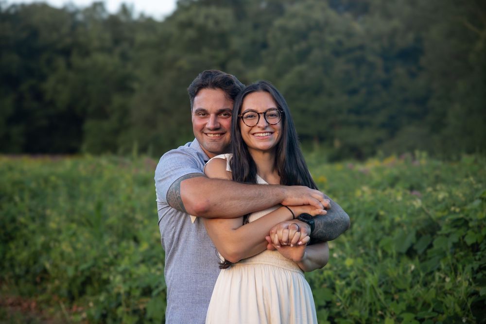 A smiling couple embraces in a lush green field, the woman wearing glasses and a white skirt.