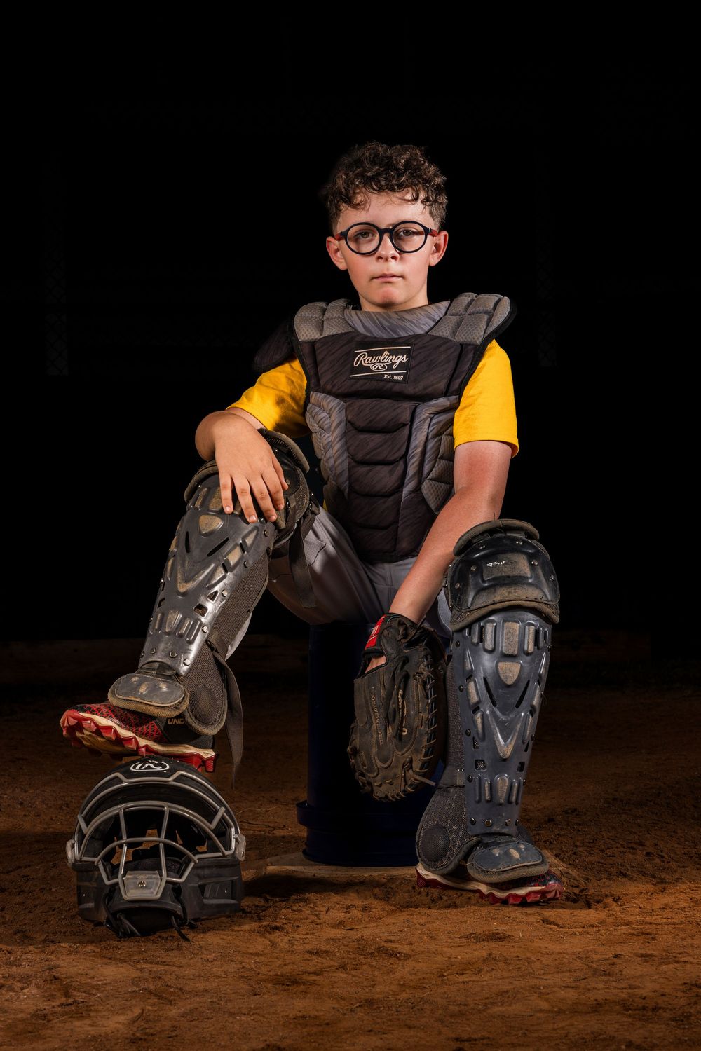 Baseball catcher's gear including chest protector, shin guards and glove arranged on dirt field against dark background.