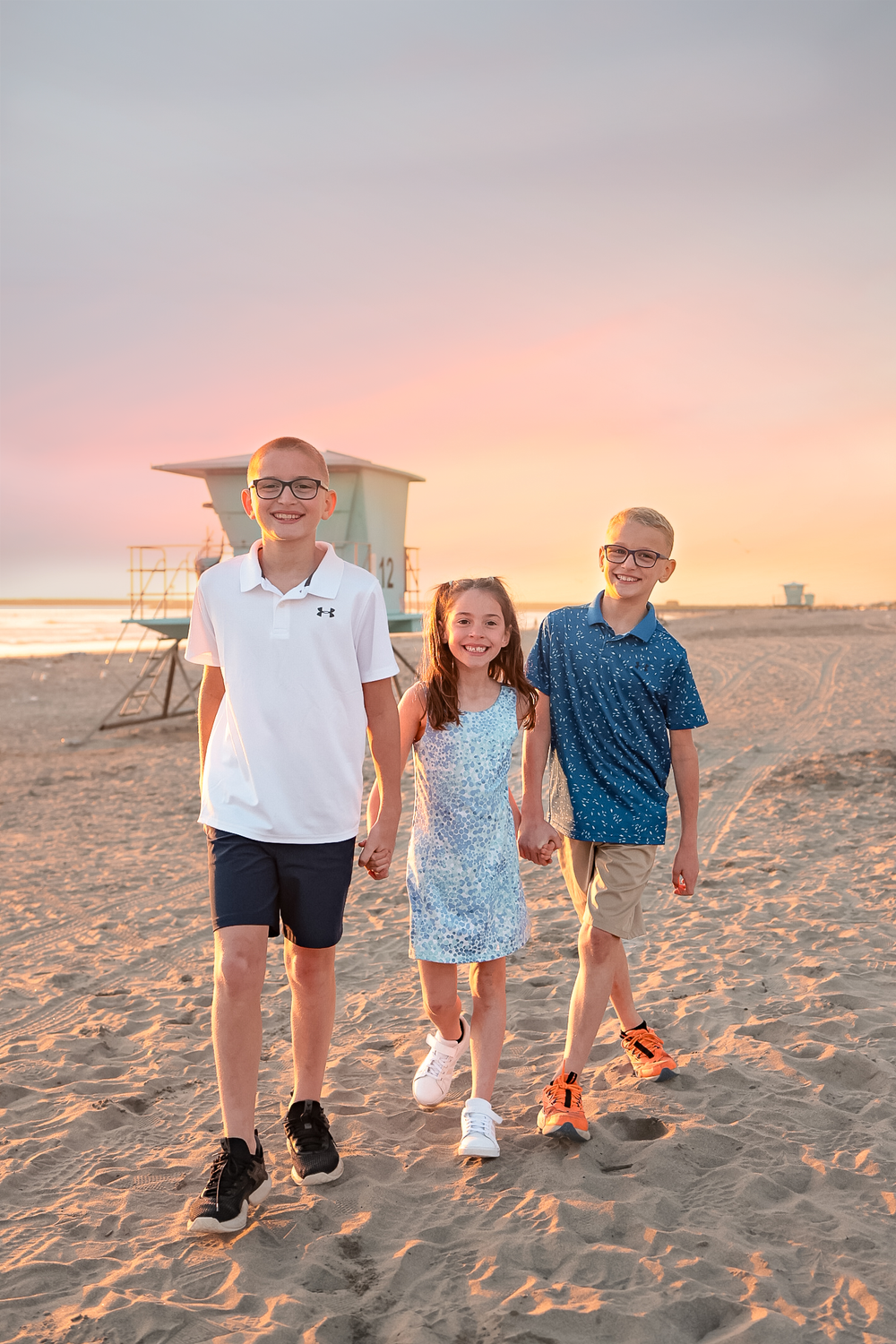 Three siblings walk together on a sandy beach at sunset with a lifeguard tower in the background.