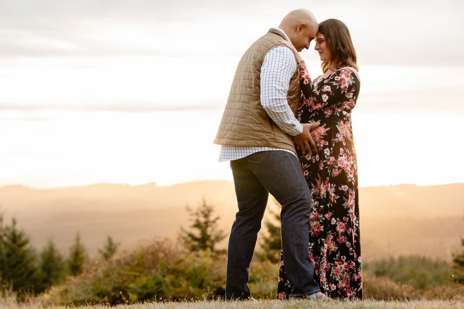 A couple embraces on a hilltop overlooking a beautiful sunset vista through evergreen trees.