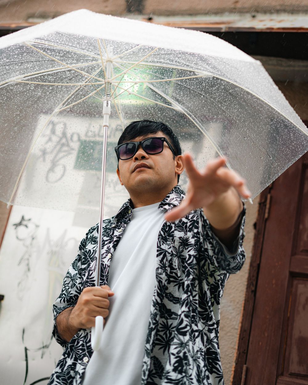 Stylish urbanite poses with clear umbrella wearing floral print shirt and sunglasses against graffiti wall.