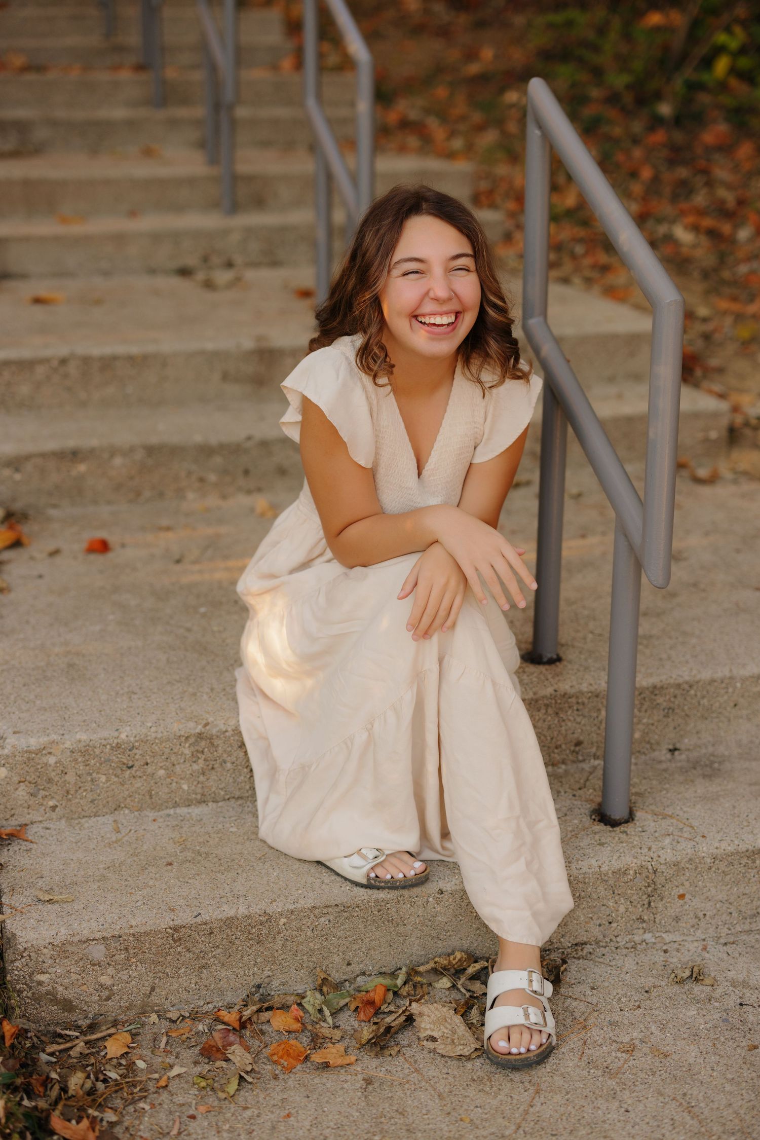 Person sits smiling on concrete steps wearing white casual dress and sandals on an autumn day with fallen leaves.