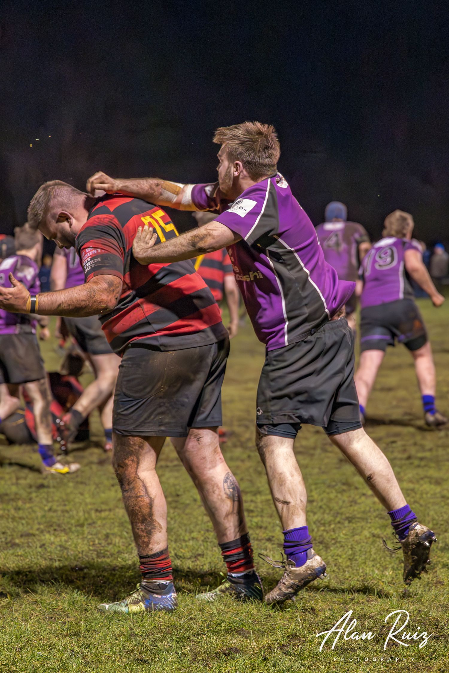 Rugby players in black and purple jerseys engage in a scrum formation during a nighttime match.