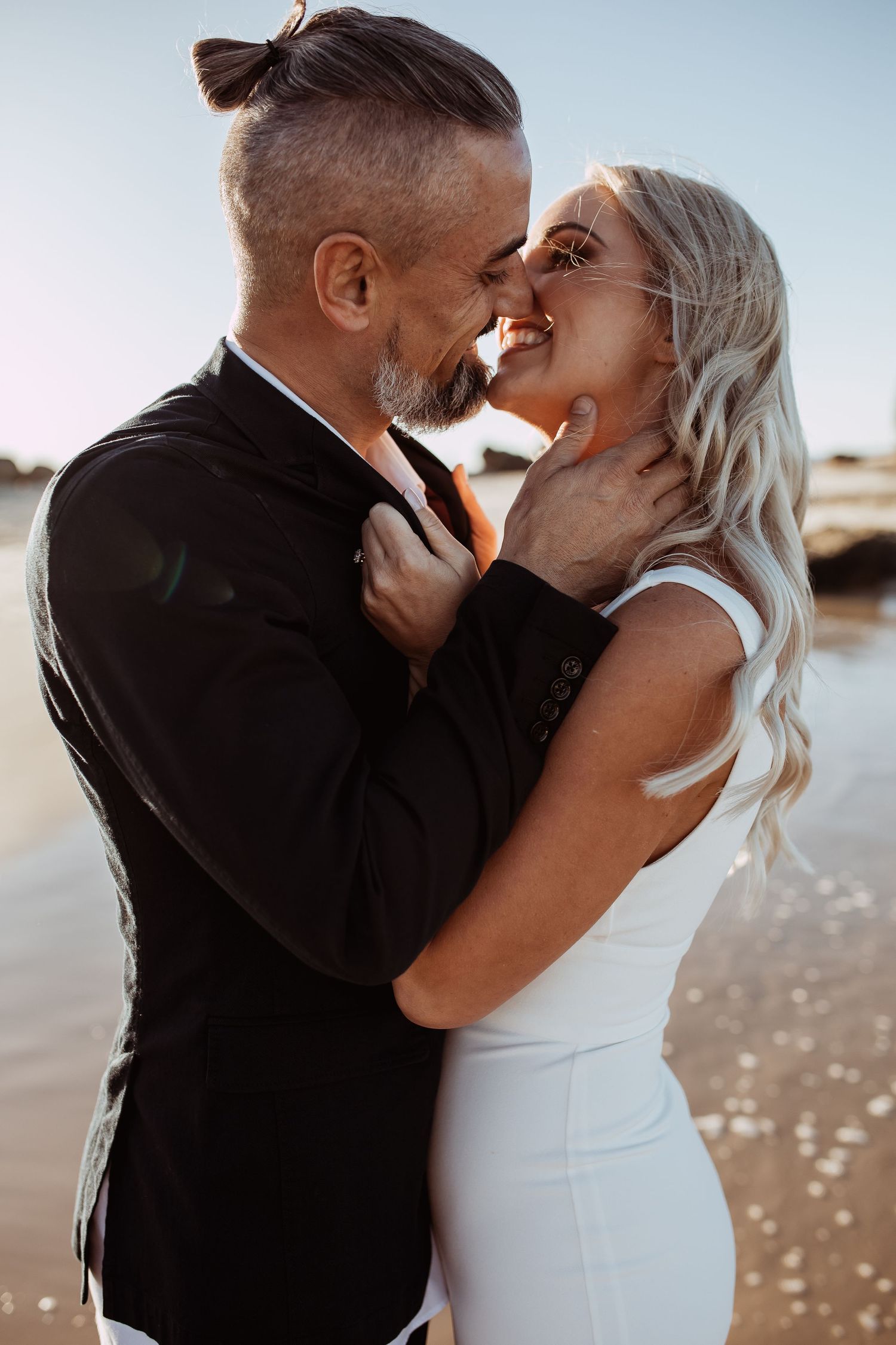 A romantic couple shares an intimate moment on the beach at sunset with her in white and him in black formal attire.