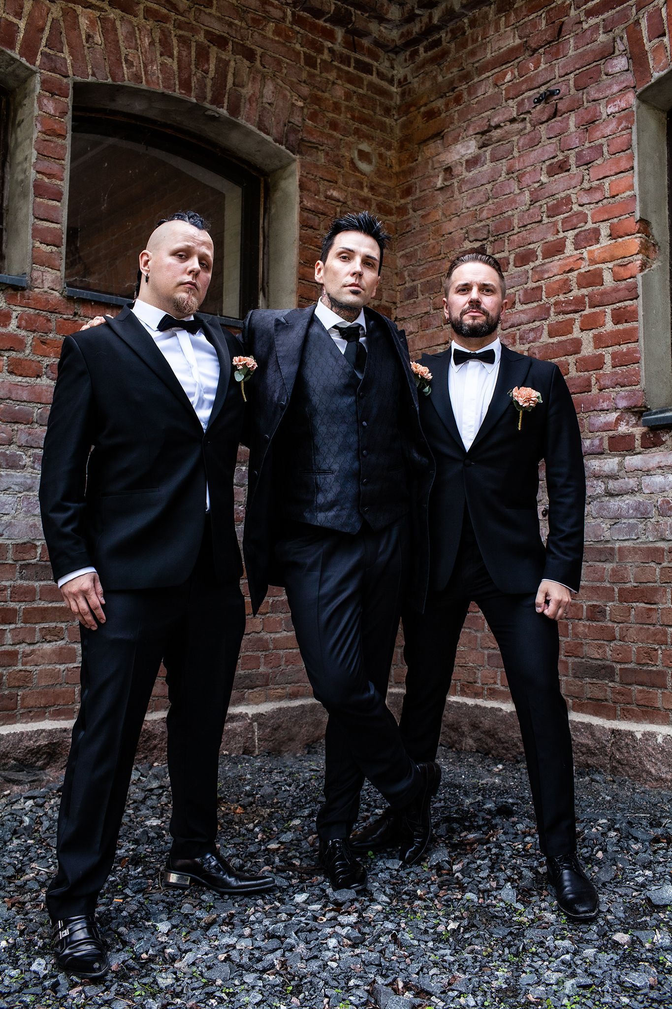 Three groomsmen in formal attire posing against a rustic brick wall backdrop.