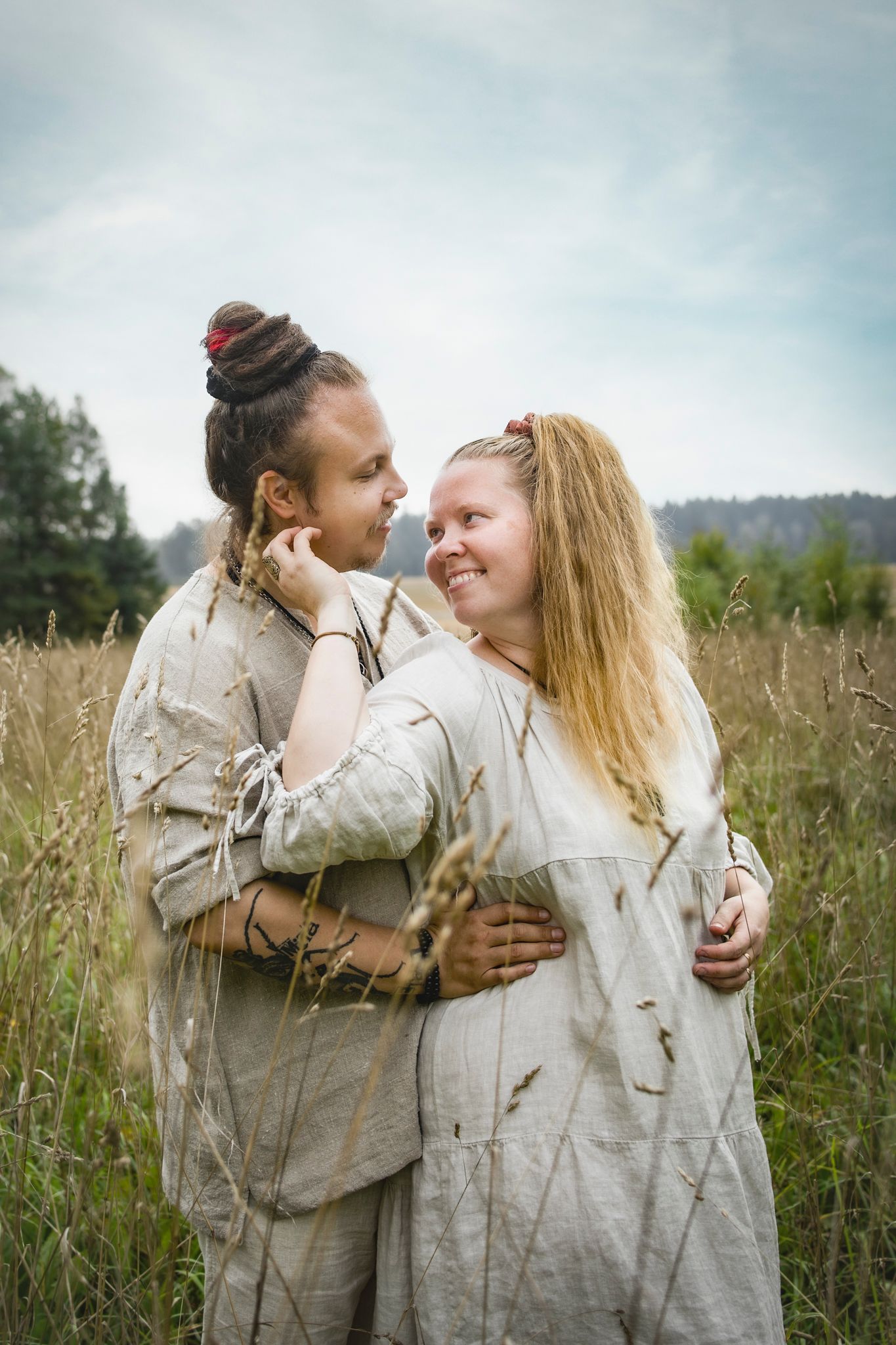 Couple in linen outfits embracing in a meadow, smiling at each other with trees and cloudy sky behind.