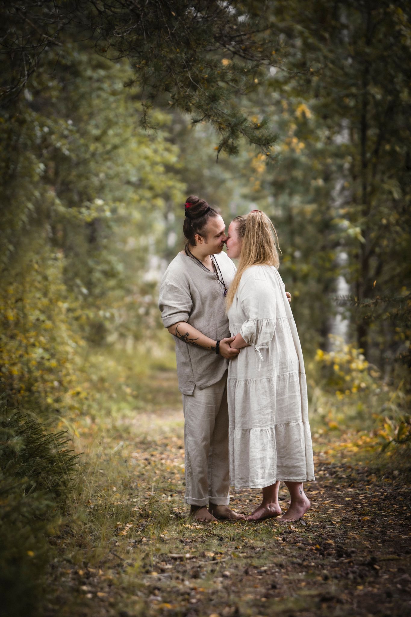 Couple kissing on a forest path, wearing matching linen outfits, surrounded by autumn foliage.