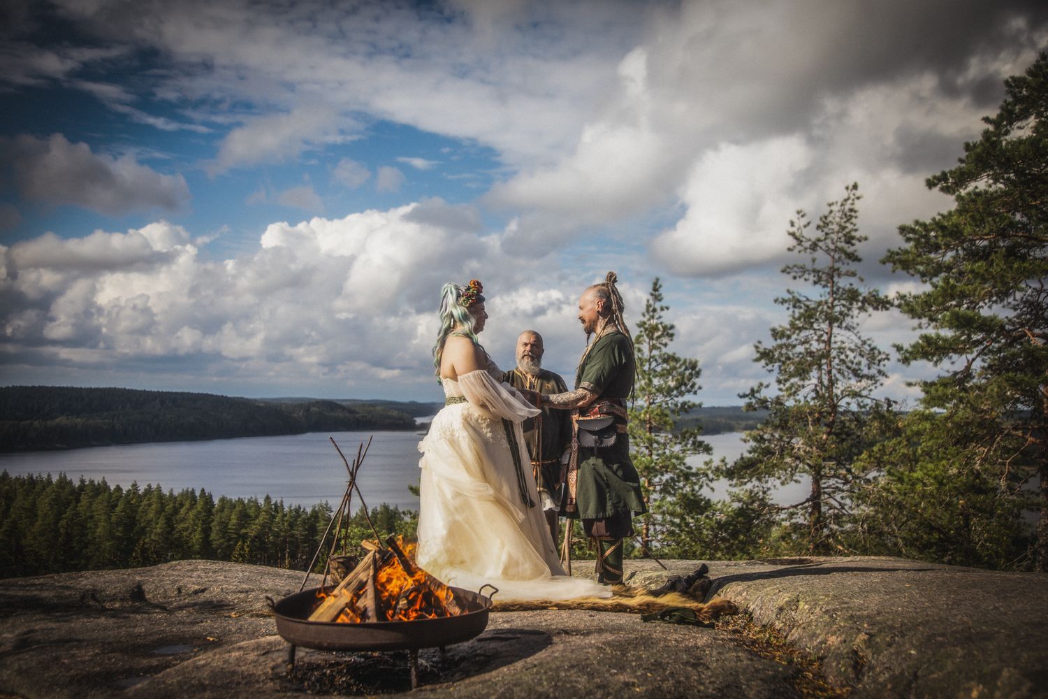 Outdoor wedding ceremony on rocky cliff with campfire, bride and groom in Celtic attire, overlooking scenic lake and forest.