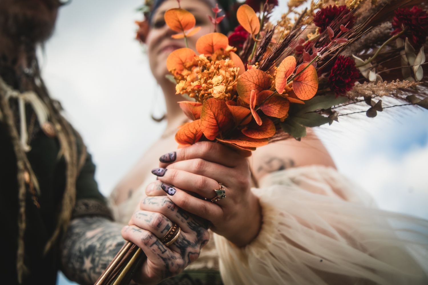 Tattooed bride holding vibrant orange and red autumn wedding bouquet with floral crown outdoors.