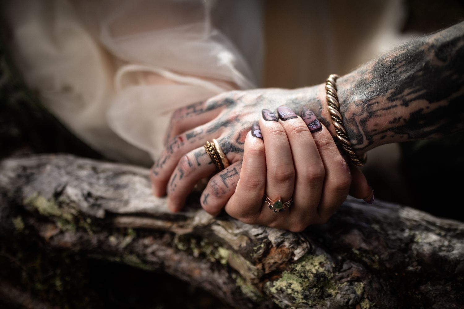 Two tattooed hands with rings and purple nails rest together on a mossy branch.