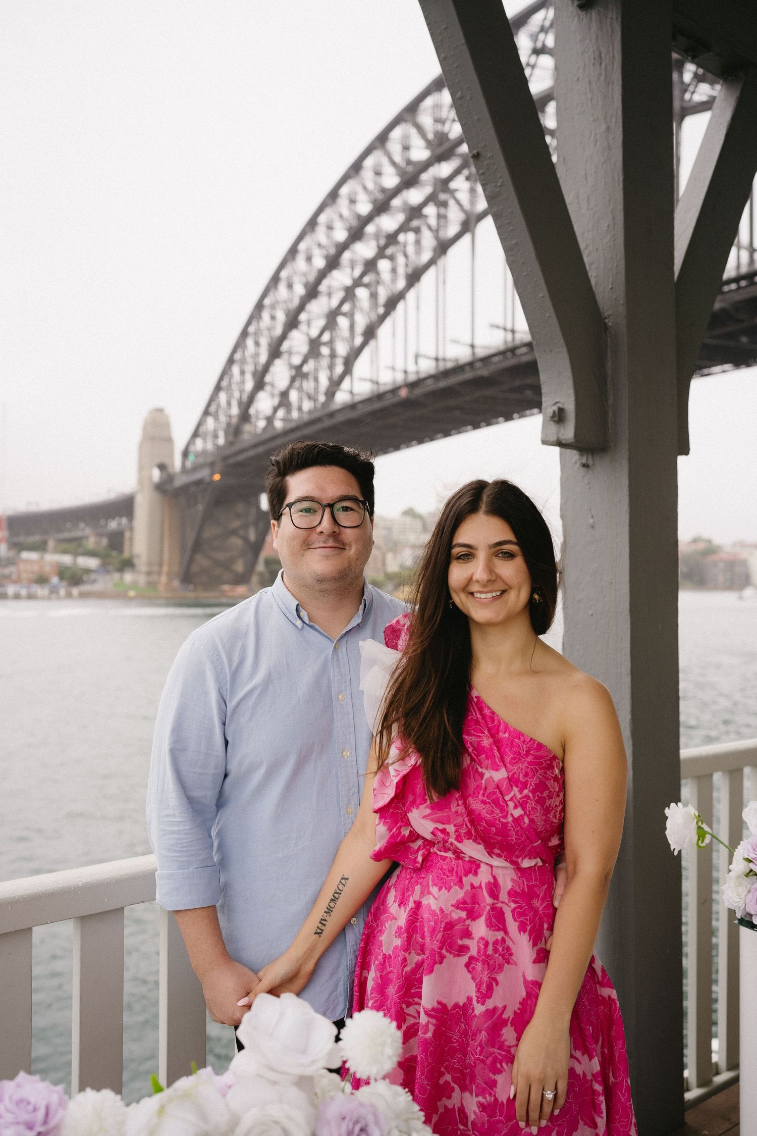 A couple poses together on a deck with the Sydney Harbour Bridge in the background.