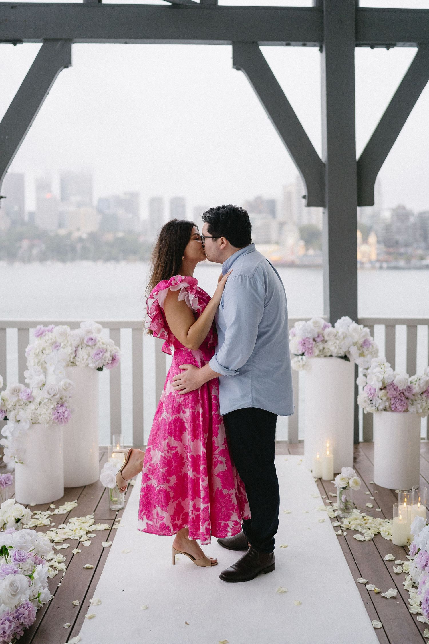 A couple shares a kiss on a decorated deck with white candles and pink flowers around them.