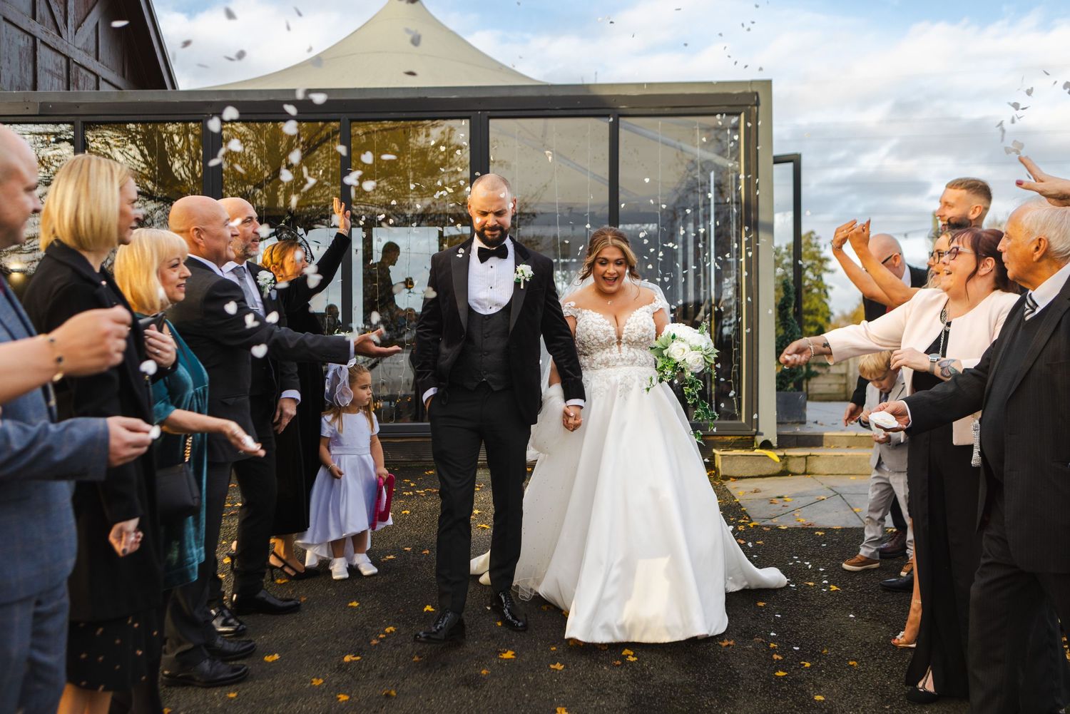 Wedding guests throwing confetti at newlyweds outside venue on autumn day.