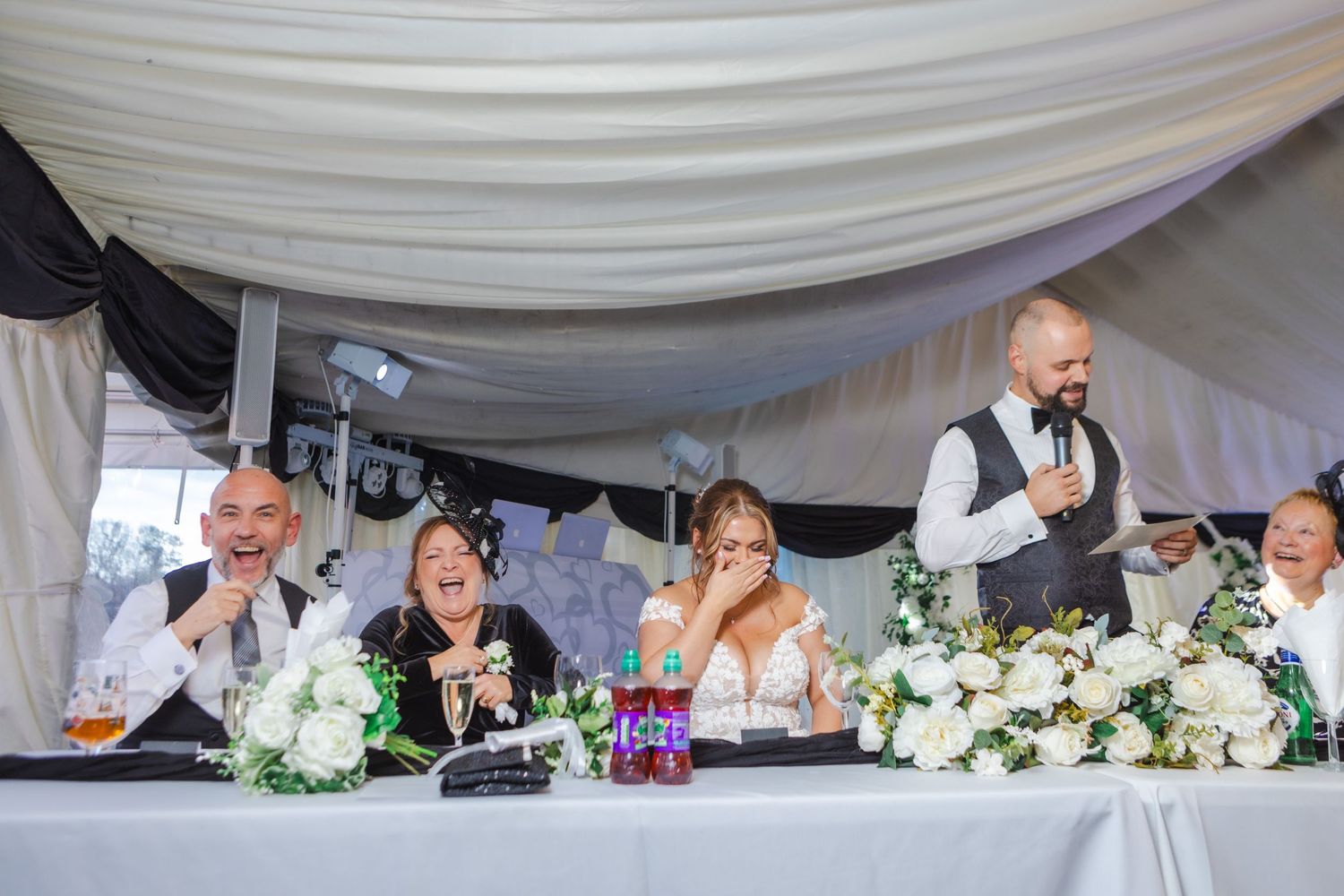 Wedding party seated at head table during reception with white floral arrangements.
