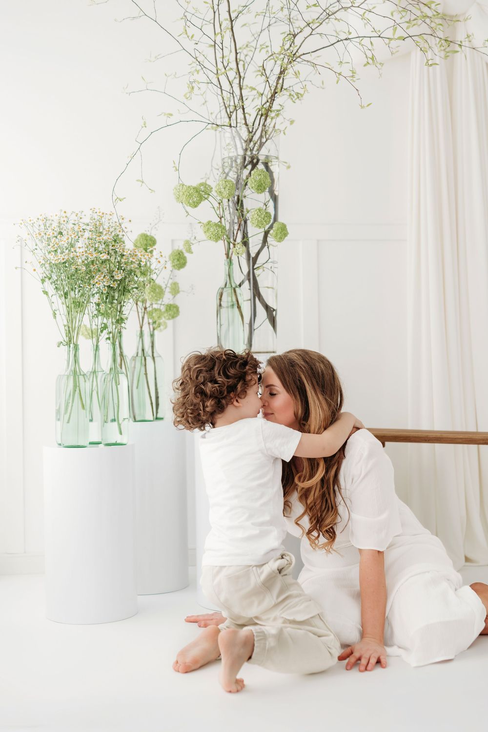 Tender moment shared in white room with green floral arrangements in glass vases on white pedestal.