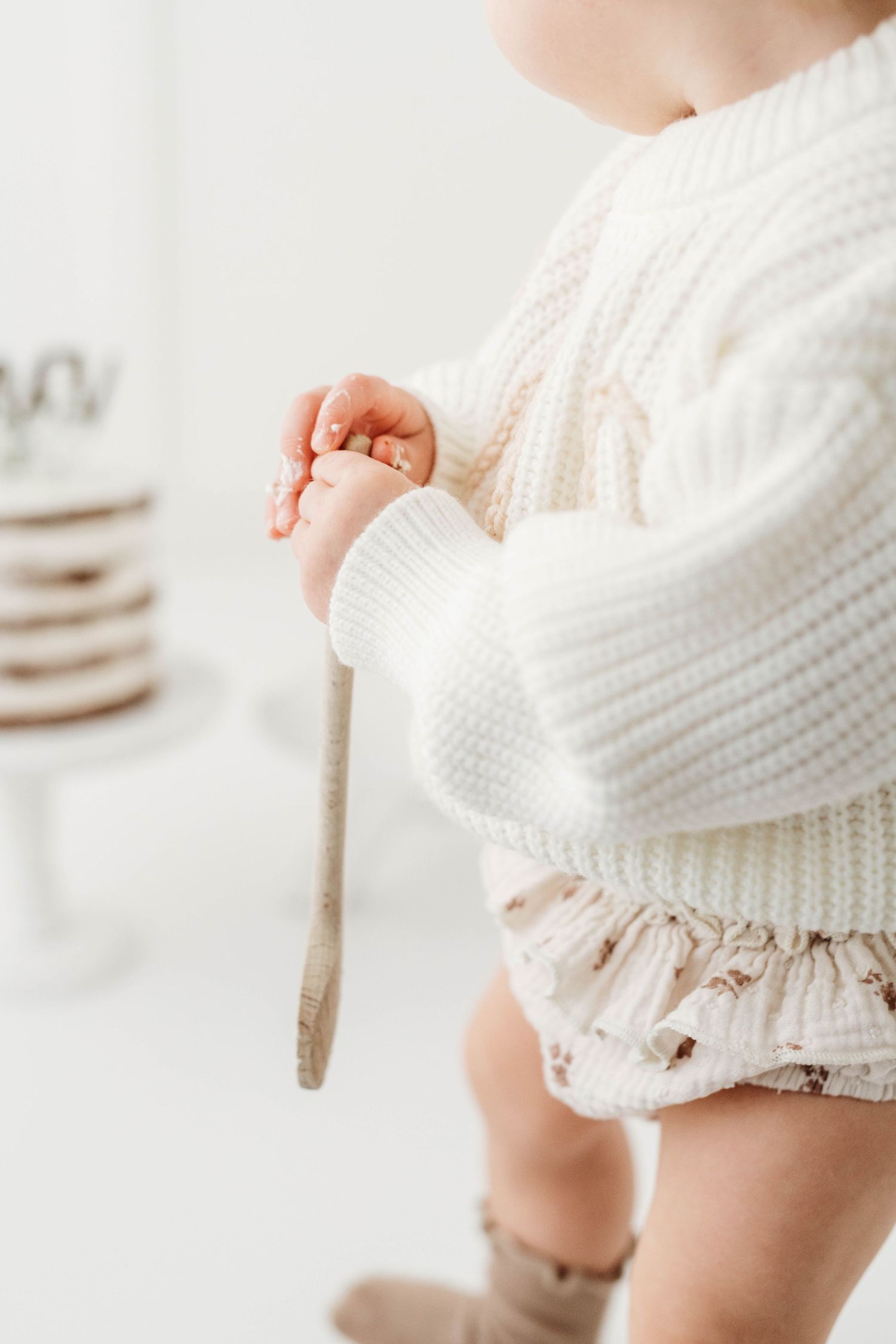 Close-up view of infant wearing white knit sweater and ruffled bloomers, holding a wooden spoon in soft lighting.