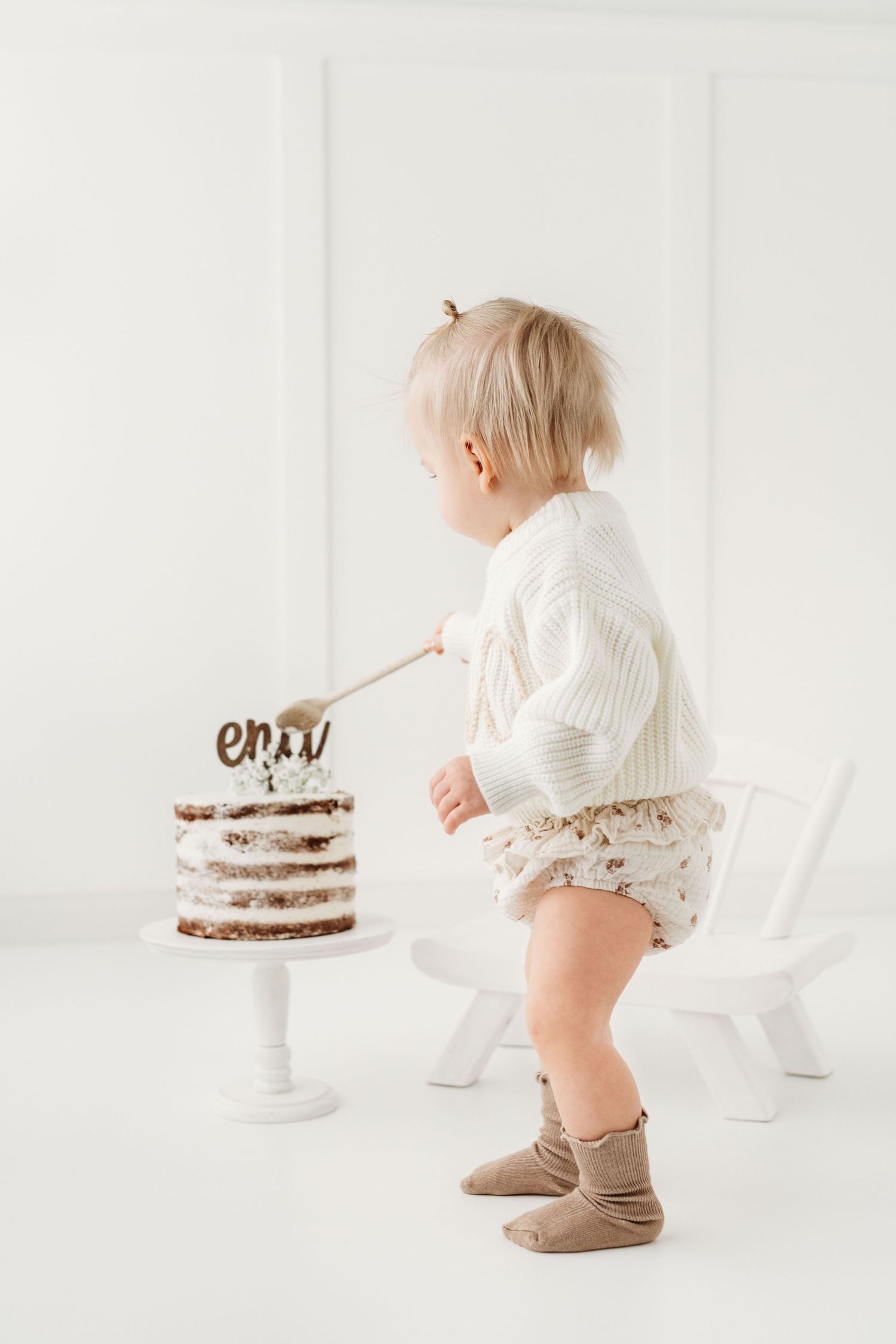Small child in white sweater and tan boots reaching toward a tiered cake on a white pedestal stand against light background.
