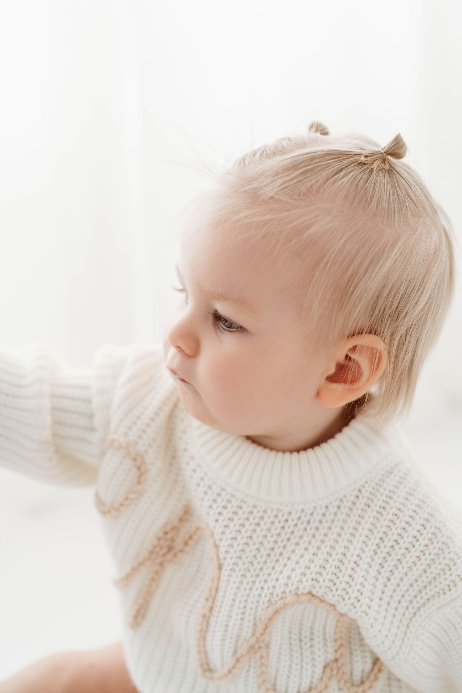Baby in white knit sweater gazes thoughtfully to the side in soft natural window light.
