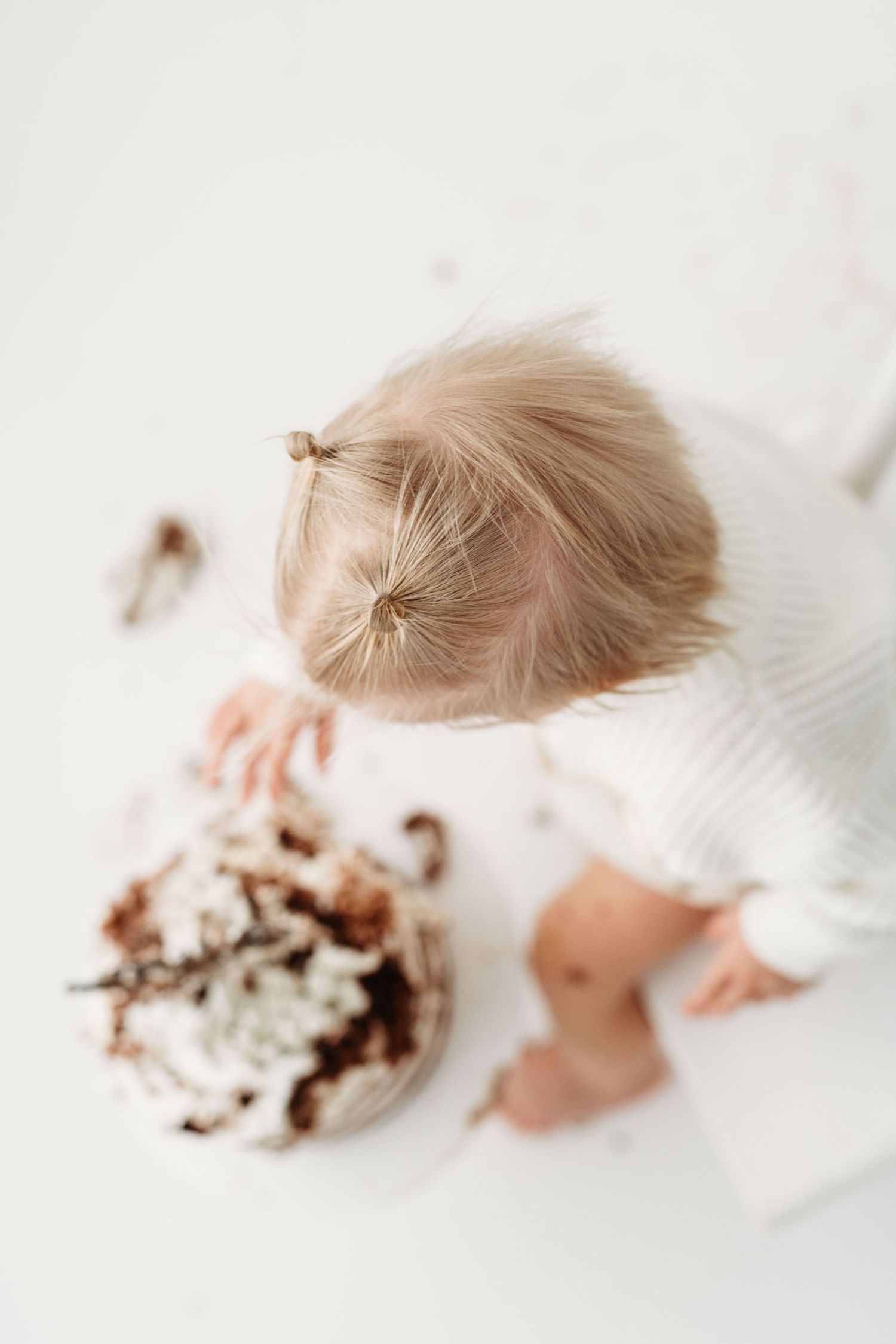 High angle view of a toddler with blonde hair sitting next to chocolate cake on white surface.