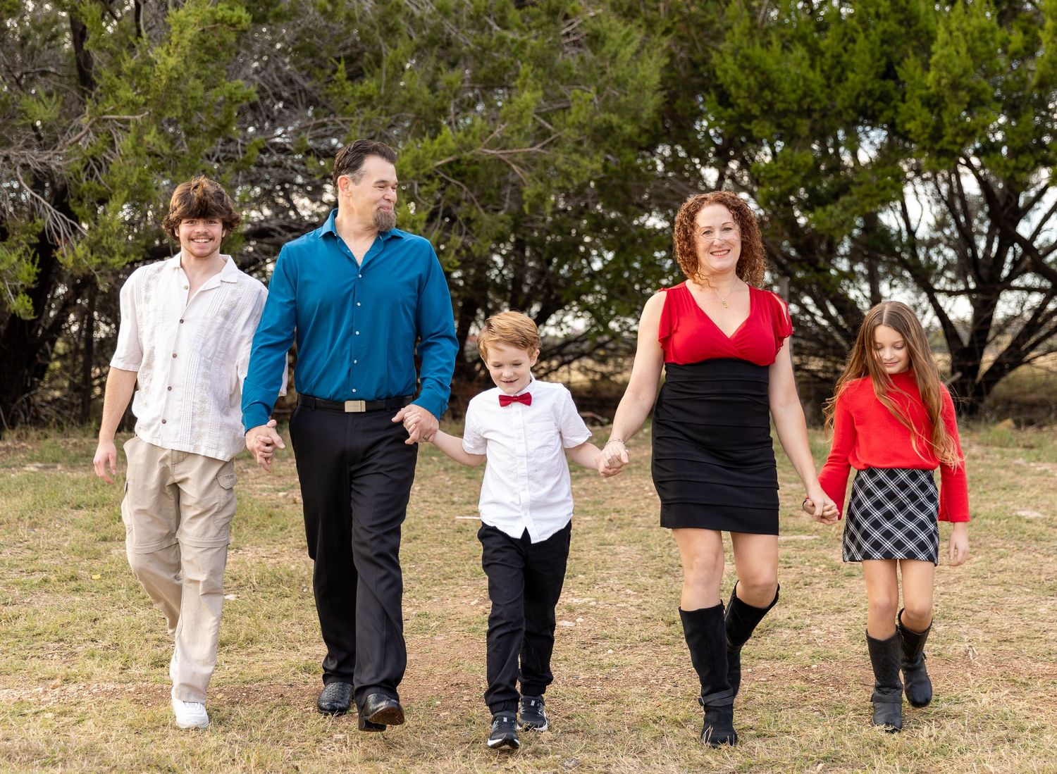 A family of five walks together holding hands on a grassy path with trees in the background.