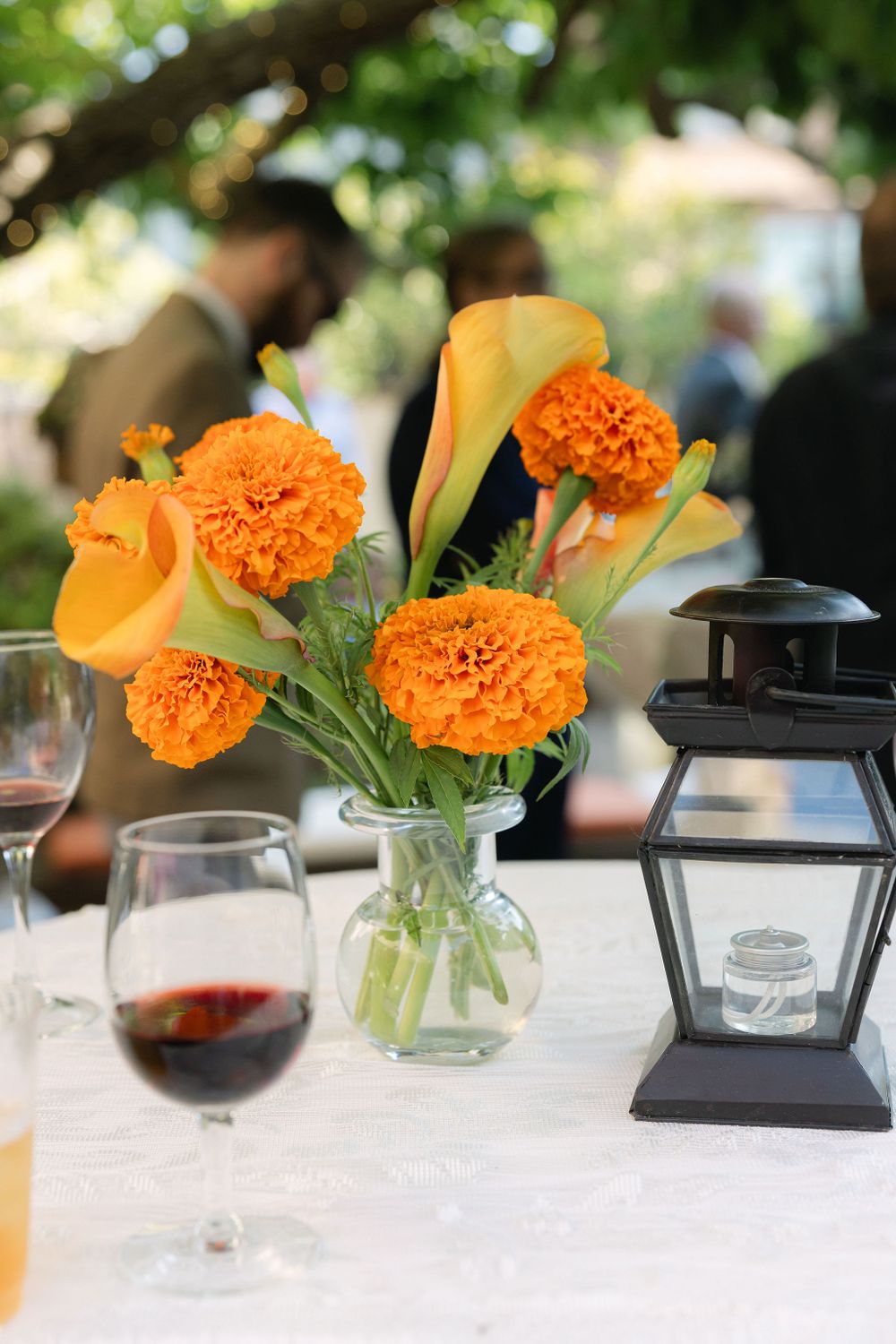 Table centerpiece with orange marigolds and calla lilies in glass vase next to black lantern and wine glass on white tablecloth.