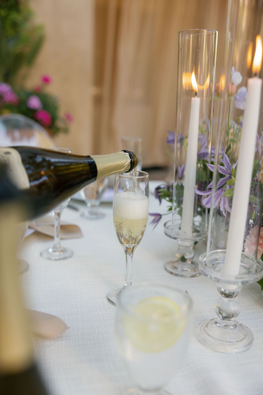 Elegant table setting with champagne glasses, tall candle holders and delicate purple flowers on white tablecloth.