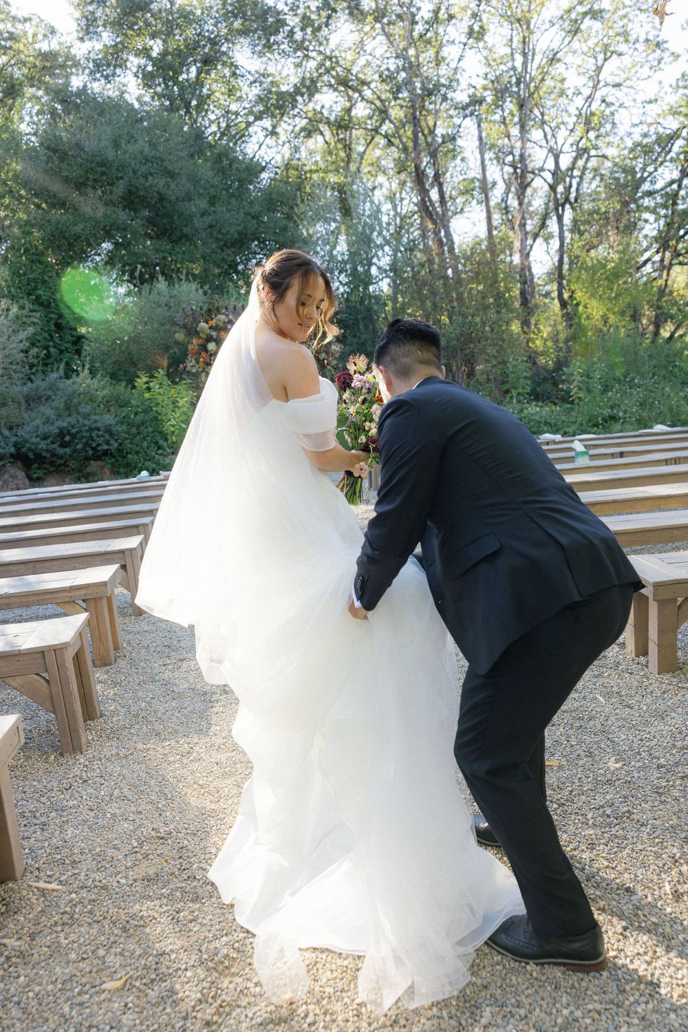 A romantic wedding moment captured outdoors with a flowing white dress and black suit at a wooded ceremony venue.