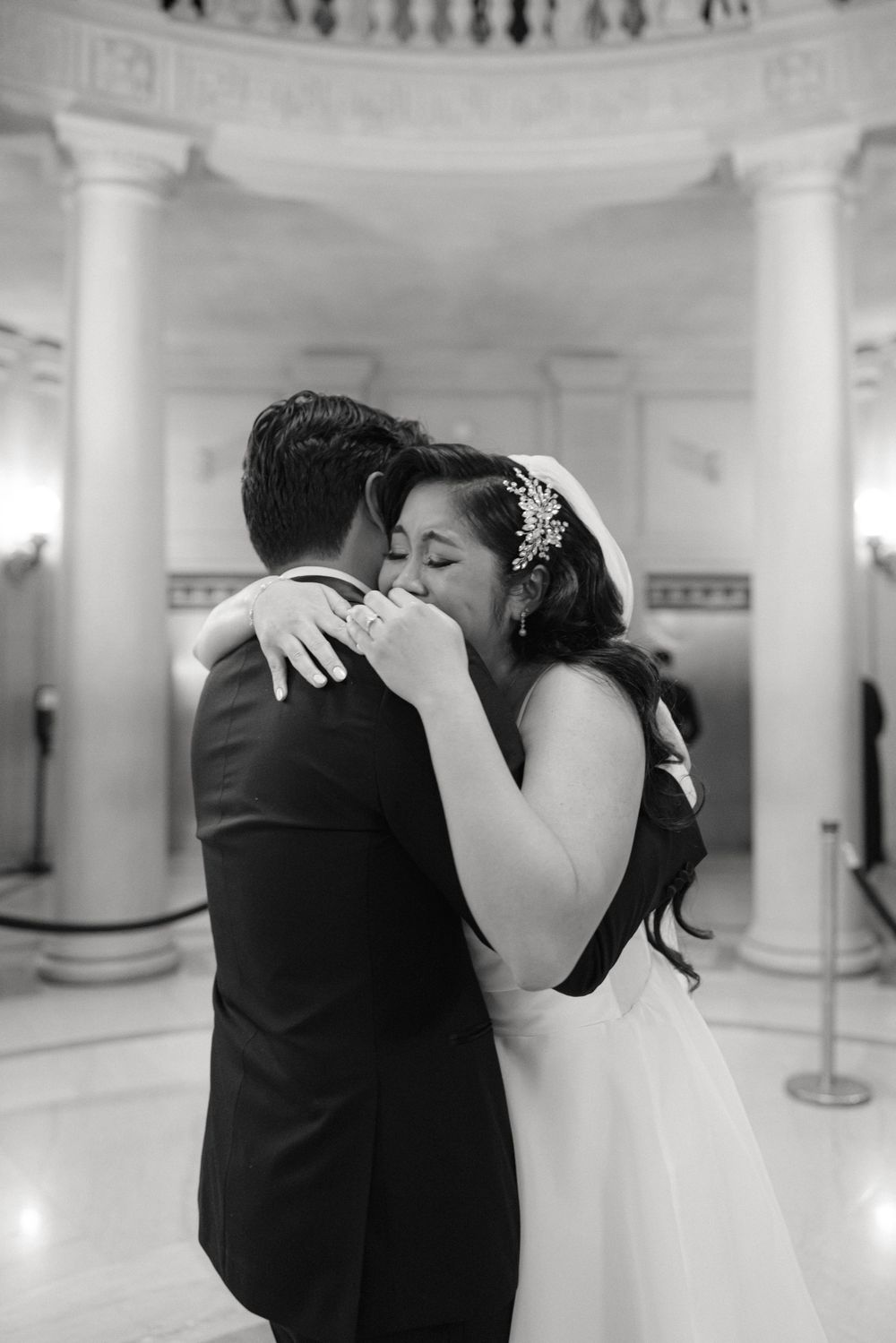 A tender black and white moment between a couple in wedding attire sharing an embrace in an elegant columned venue.