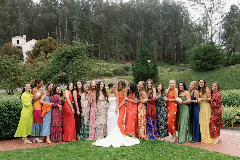 A large group of attendees in colorful formal dresses stand together at an outdoor garden wedding ceremony.
