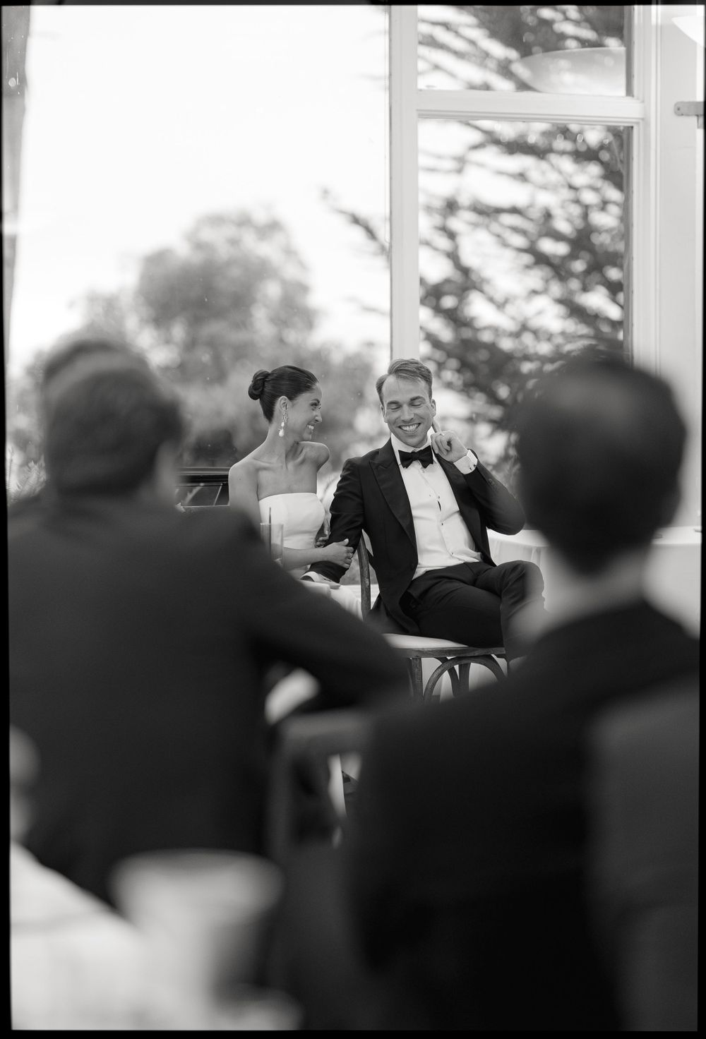 Black and white photo of a wedding reception showing a couple seated and laughing during speeches at an indoor venue.