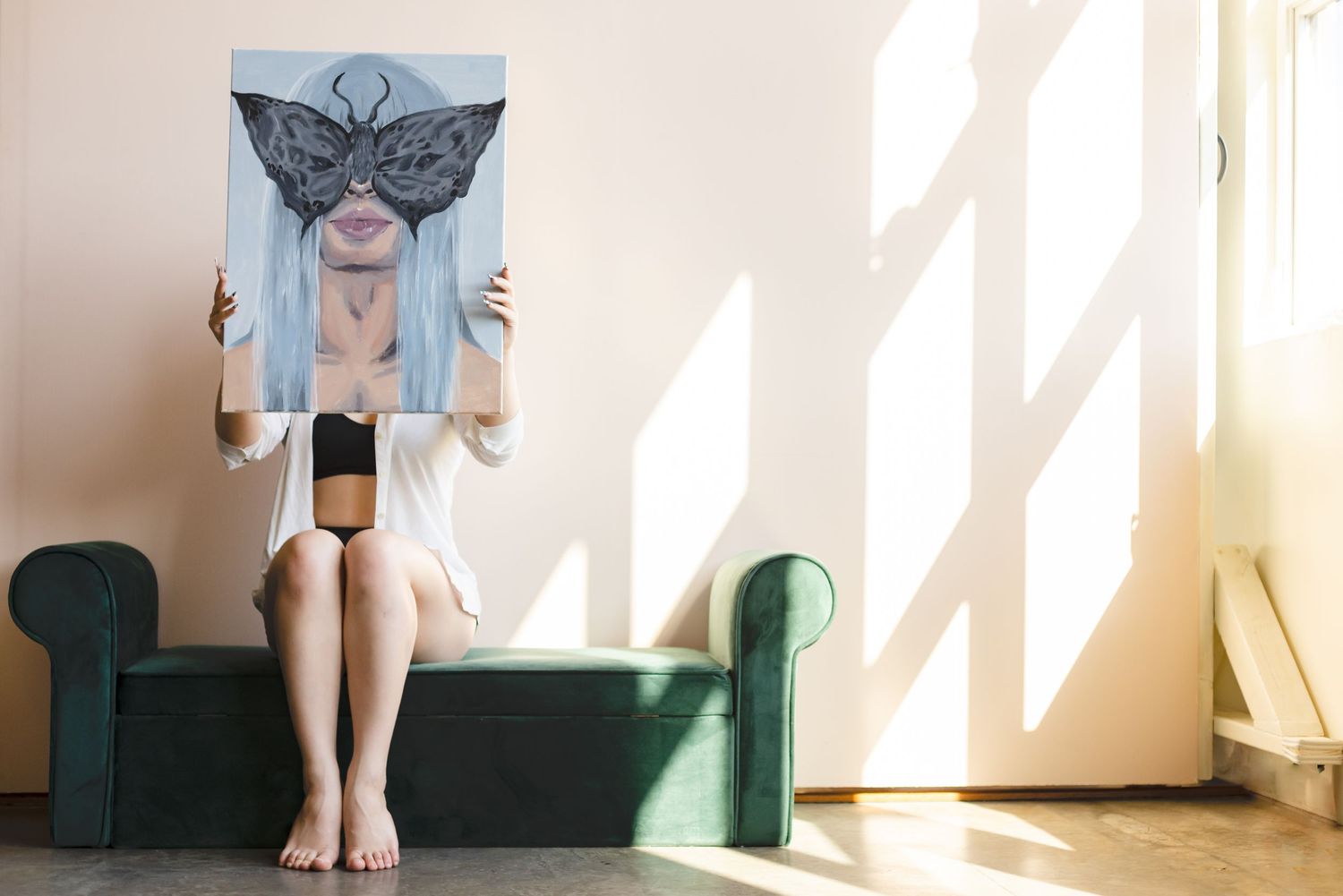 Person sitting on green couch holding up a drawing of butterfly face in front of their face in sunlit room.
