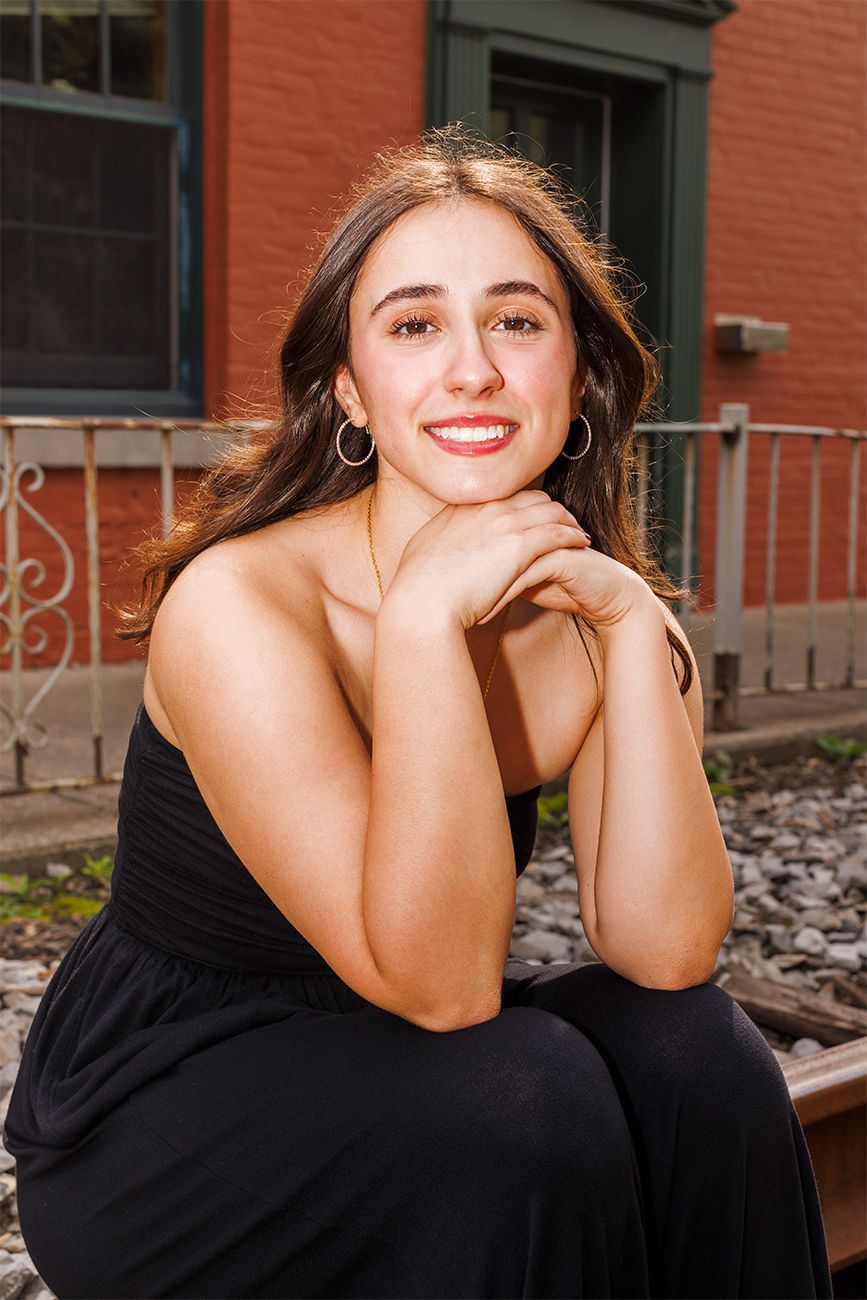 Portrait of a person in a black dress sitting outdoors near a brick building.